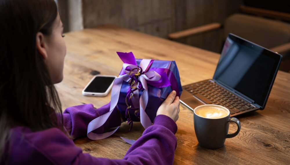 Young woman in a purple sweater with a purple gift box in a cafe with a cup of coffee in front of a laptop.